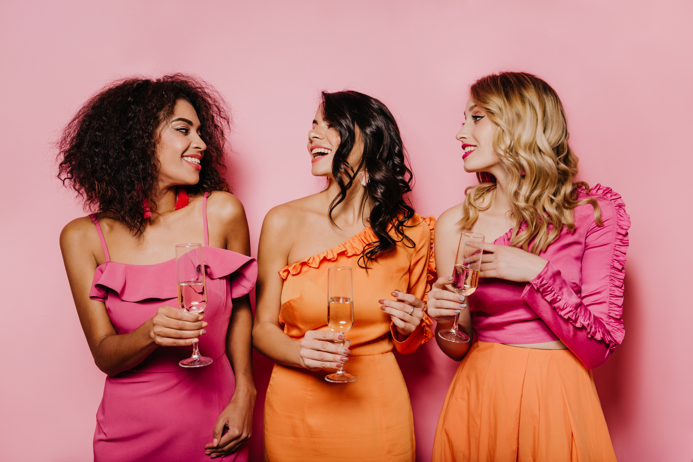 Pleased Girls Talking and Drinking Champagne. Well-Dressed Ladies Posing on Pink Background with Wineglasses